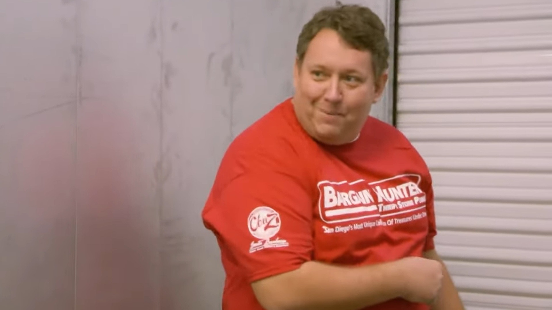 Storage War's Rene Nezhoda pointing towards a storage lot door. He is wearing a red t-shirt that says 'Bargain Hunter' in white text. 