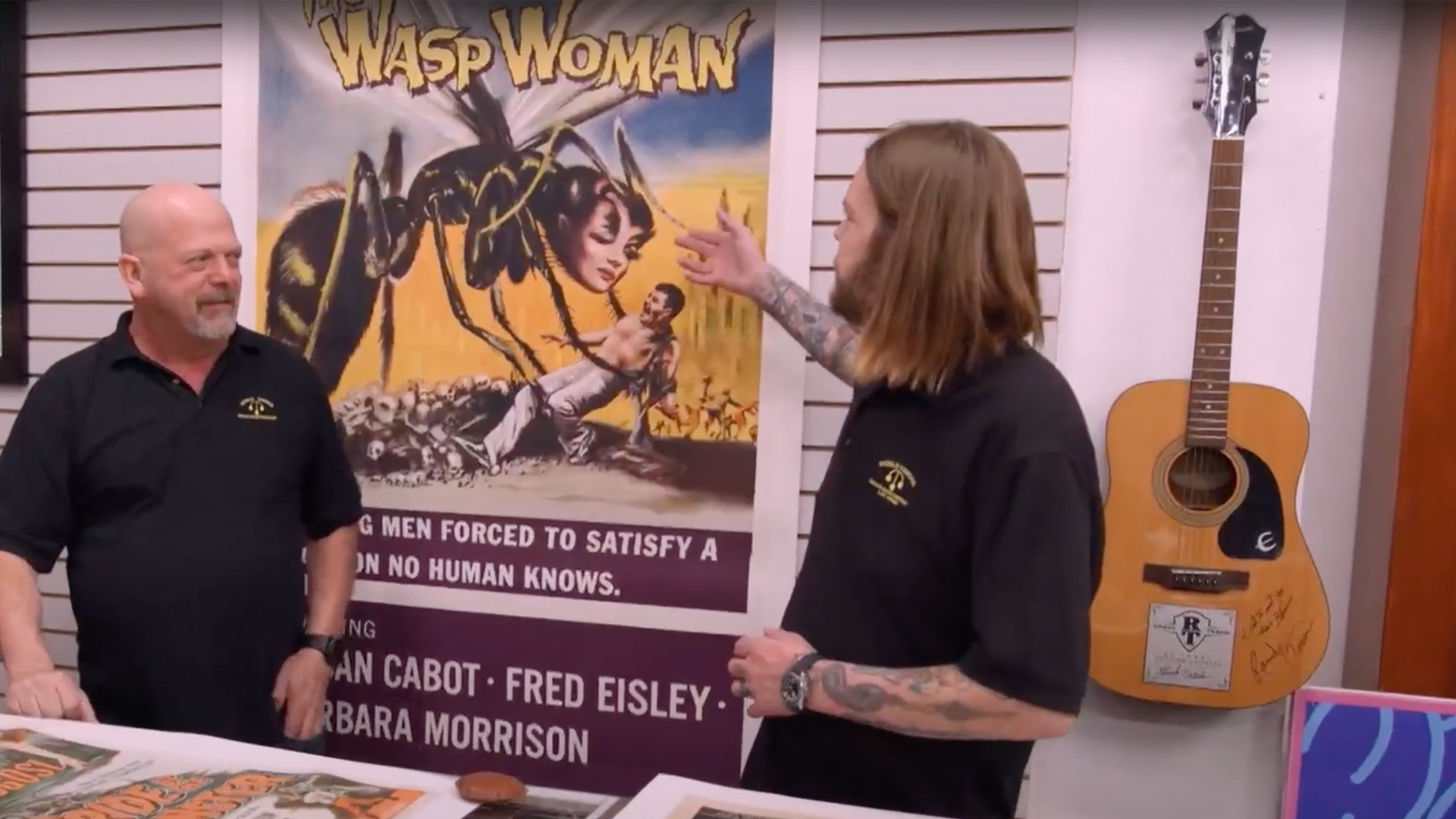 Rick and Corey Harrison stand in front of a large poster for an old movie titled 'The Wasp Woman'