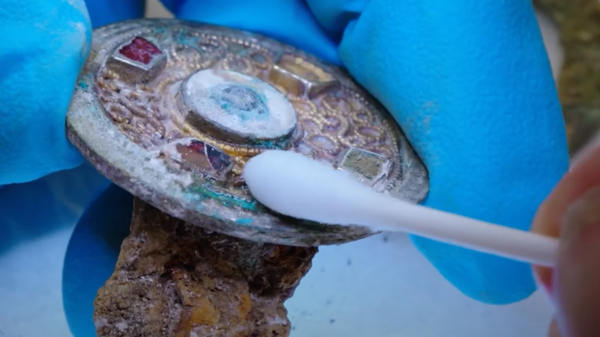 An archaeologist uses a cotton bud to gently clean an old jeweled brooch that had been found in a field