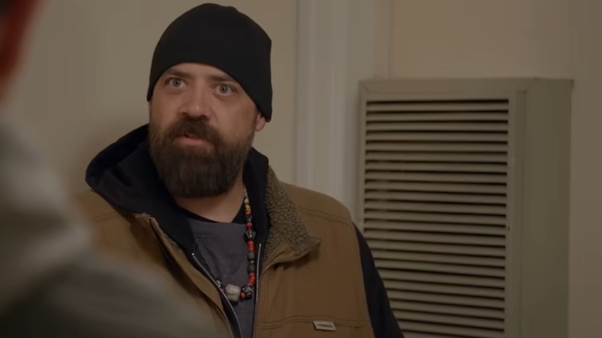 A bearded man wearing a black beanie and brown jacket with a fleece collar stands indoors near a wall vent, looking intently at someone off camera.