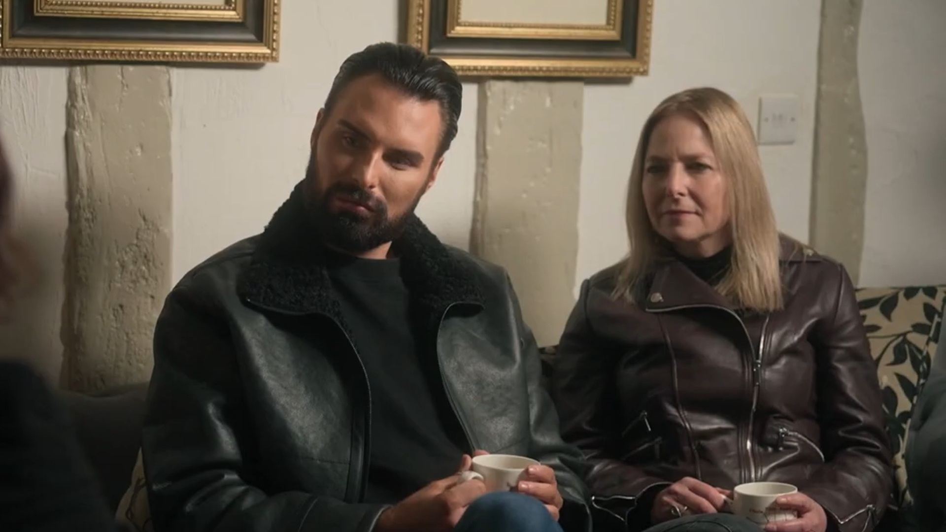 Rylan Clark and Professor Alice Roberts on Witches of Essex. They are seen sitting together in a house holding cups of tea and looking intently at someone speaking off screen.
