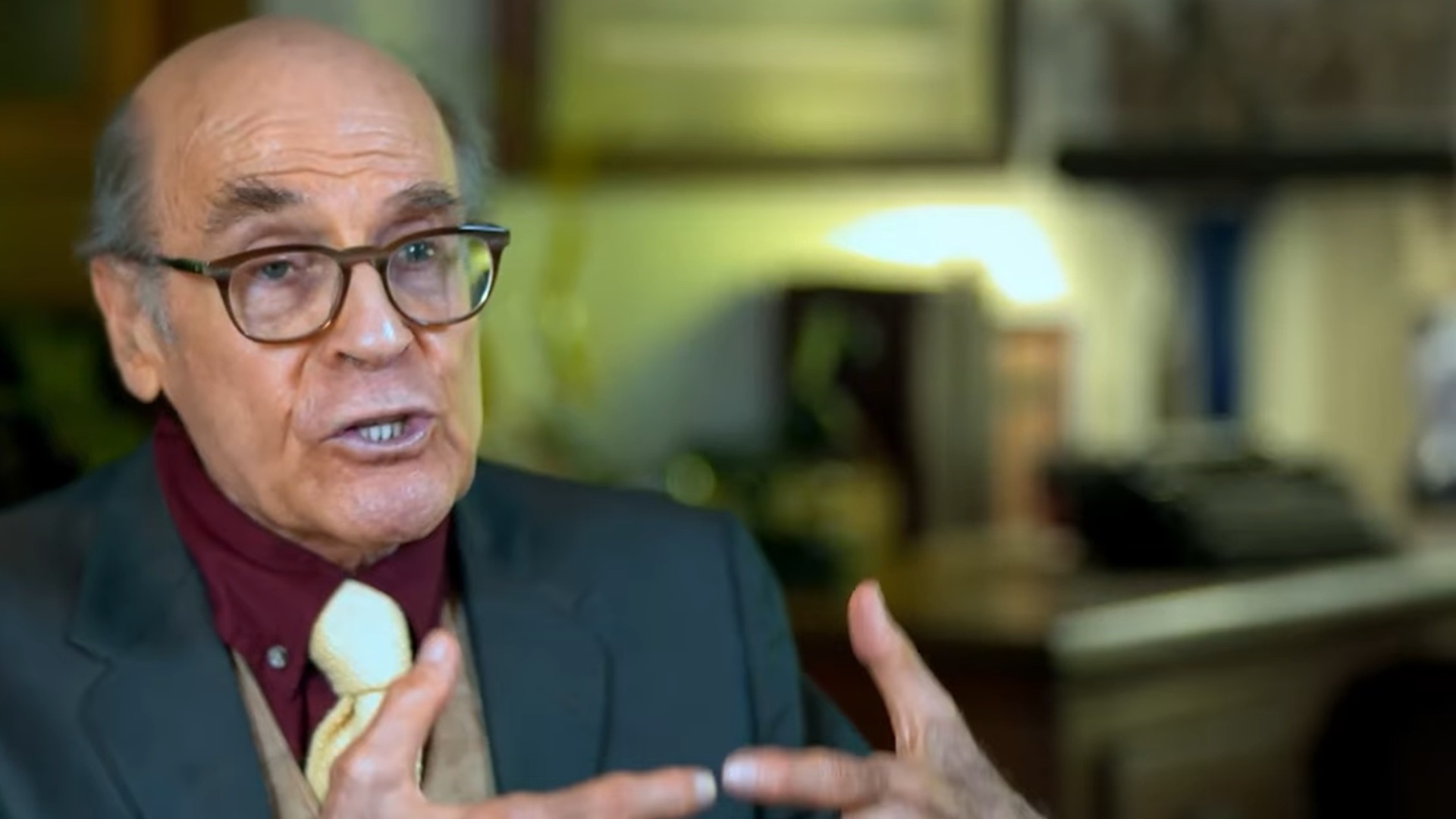 An older man wearing glasses, a dark suit, a maroon shirt, and a light yellow tie speaks while gesturing with his hands in a warmly lit room.