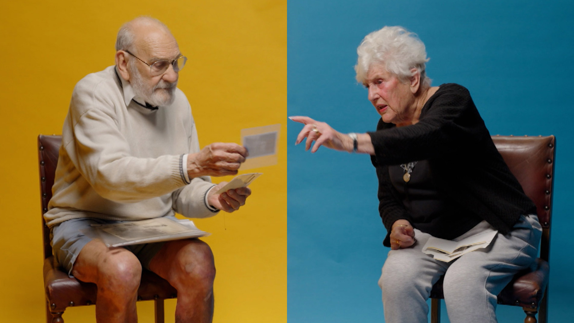 Elderly man and woman sit on chairs against yellow and blue backdrops, sharing and discussing old photos