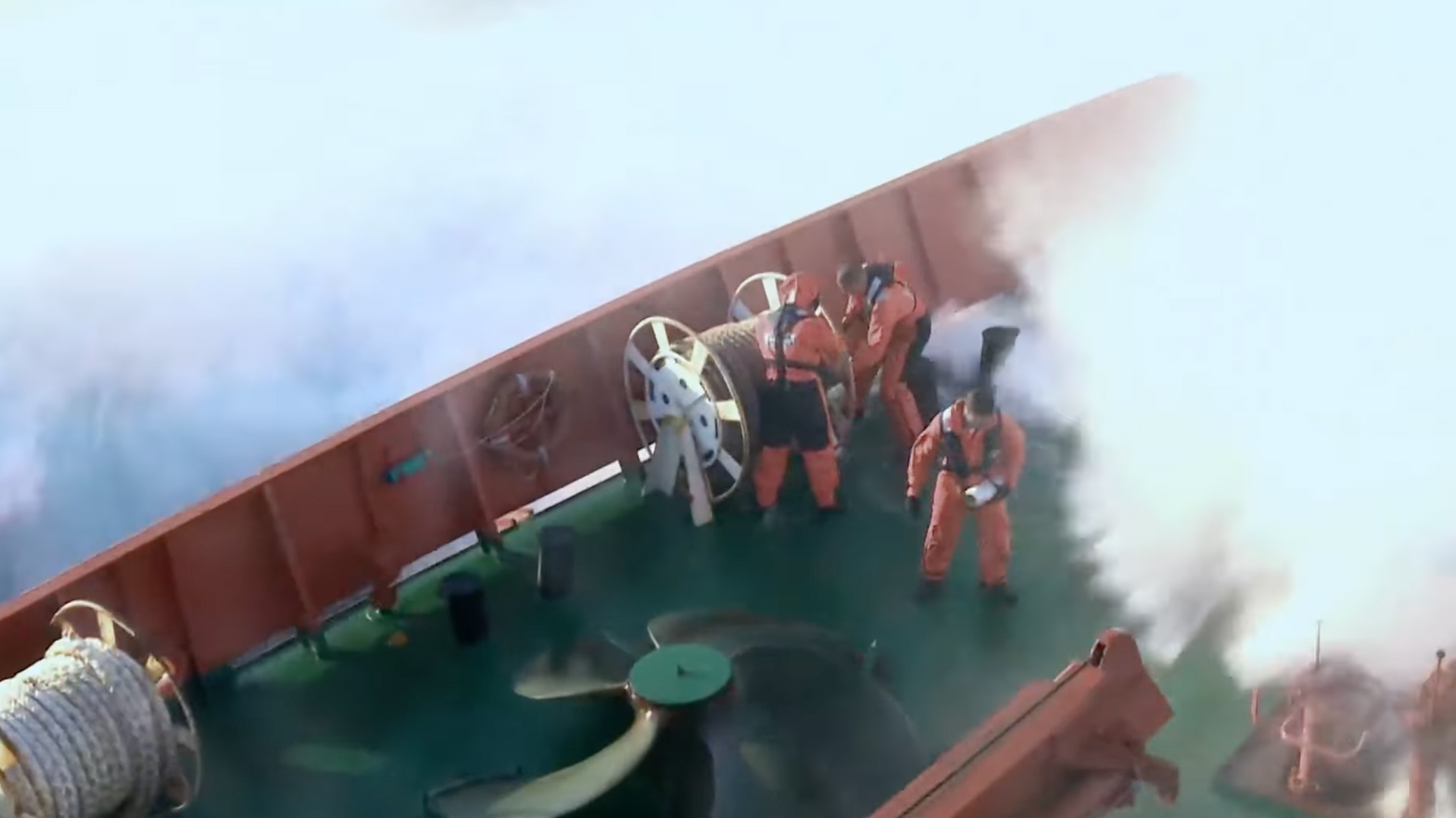 Crew members in protective gear work on a ship’s deck amid heavy spray and rough seas