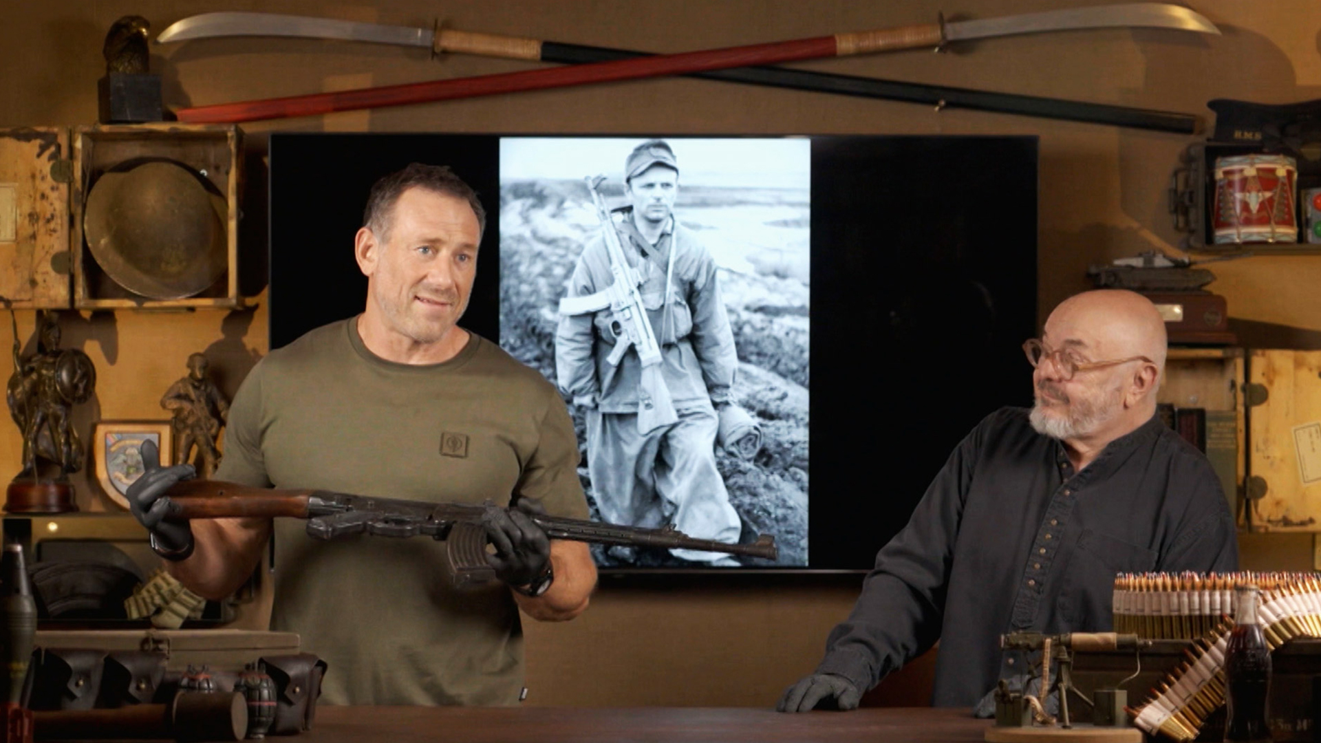 Jason Fox holding a large gun while Bruce Crompton looks on