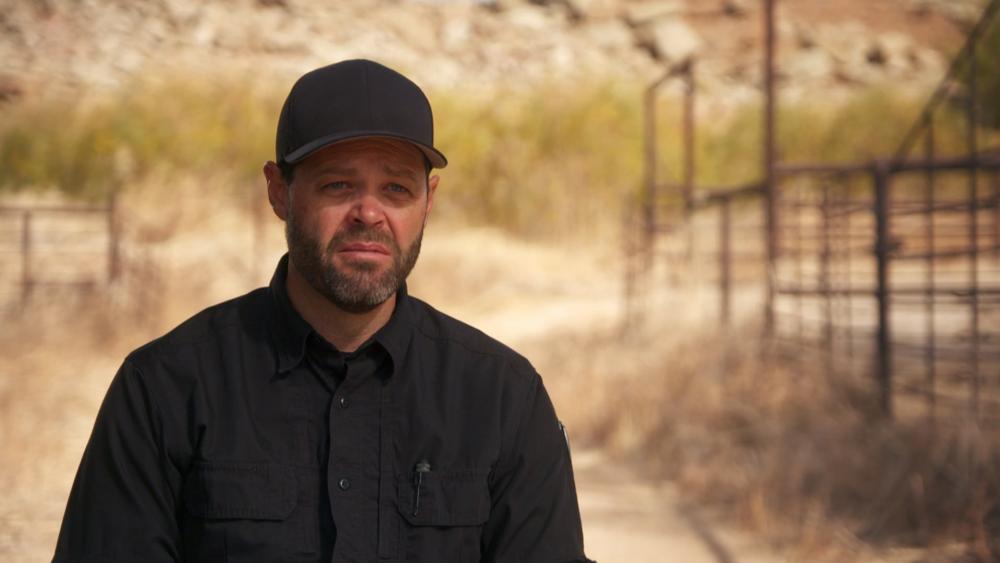Bryant 'Dragon' Arnold is wearing a black baseball cap and a black polo shirt with ranch-style fencing in the background. He is about to talk to the camera