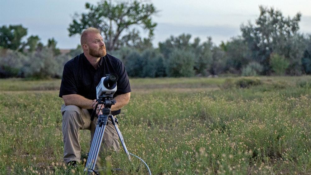 Kaleb Bench looks to the sky while on one knee in a grassy field. A high-tech camera is on a tripod in front of him and is pointing to the sky in the same direction that Kaleb is looking