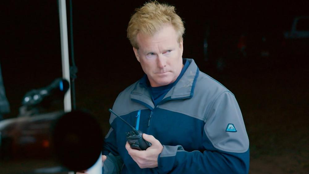 Travis Taylor looks stern while holding a walkie-talkie and wearing a fleece