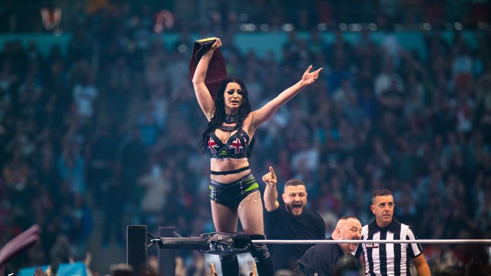 Paige holds a belt above her head while she stands on the ropes of wrestling ring