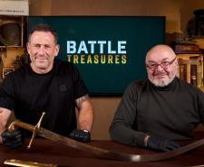 Jason Fox (L) and Bruce Crompton (R) sit in front of a television displaying the 'Battle Treasures' logo and with a large sword in front of them