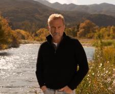 Kevin Costner stands next to a stream in the middle of rural America