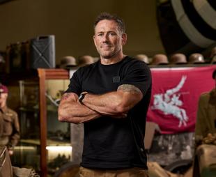 Jason Fox standing with his arms crossed in front of a lot of military artefacts