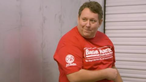 Storage War's Rene Nezhoda pointing towards a storage lot door. He is wearing a red t-shirt that says 'Bargain Hunter' in white text. 
