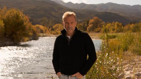 Kevin Costner stands next to a stream in the middle of rural America
