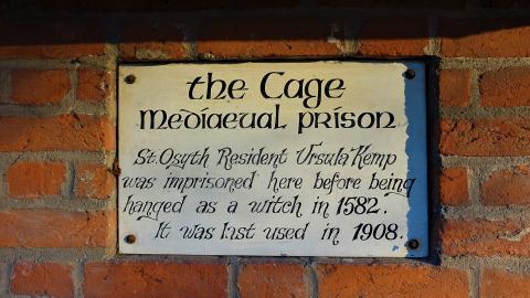 A plaque on the side of a brick building that reads, 'the Cage mediaeval prison  St. Osyth Resident Ursula Kemp was imprisoned here before being hanged as a witch in 1582. It was last used in 1908.'
