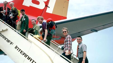 A photograph of seven men of different ages climbing the steps on to a passenger jet. They are seen looking in the direction of the photographer. The red and gold colours of the tailplane is visible, as are the words 'Braniff International' on the steps.