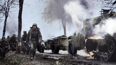 Colourised image of German soldiers passing burning U.S. equipment in the beginning of the Battle of the Bulge