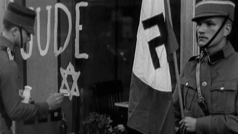 A Nazi soldier holds a swastika flag while another paints the Star of David on a shop window