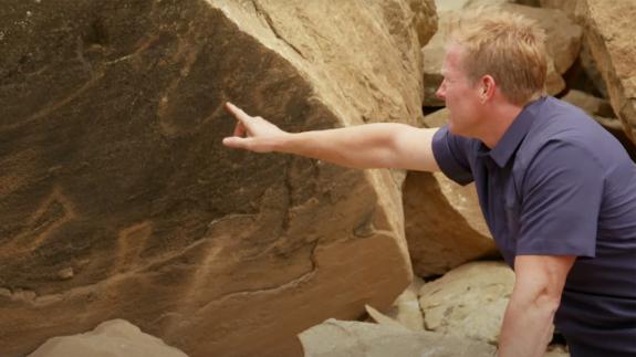 Travis Taylor points at strange lines and marking on the side of a boulder
