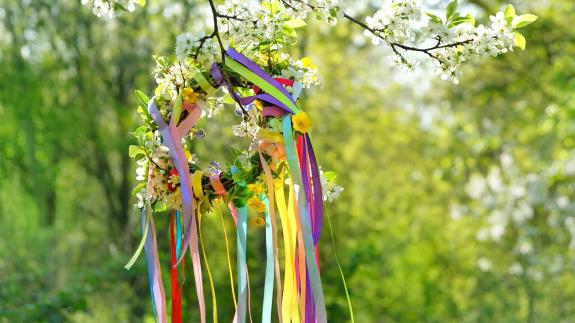 Spring flower wreath with colorful ribbons on tree in garden, green natural background. floral decor. Symbol of Beltane, Wiccan Celtic Holiday beginning of summer. pagan witch traditions, rituals flower wreath with colorful ribbons on tree in garden, green natural background. floral decor. Symbol of Beltane, Wiccan Celtic Holiday beginning of summer. pagan witch traditions, rituals