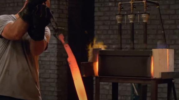 A man dips a glowing hot sword straight from the forge into a barrel of water