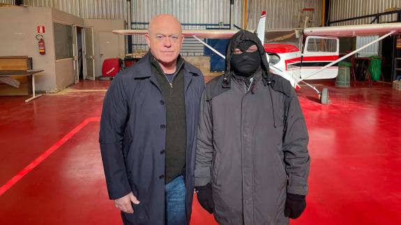 Ross Kemp stands next to a masked man in an aircraft hangar 