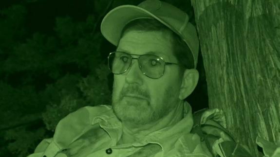 A veterinary pathologist wearing camouflage clothing sitting besides a tree trunk in the dark. He is wearing a cap, and has a pensive expression on his face.