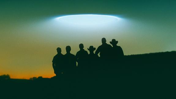 Silhouette of the Curse of Skinwalker Ranch team against a hazy evening sky with lights from a UFO hovering above them