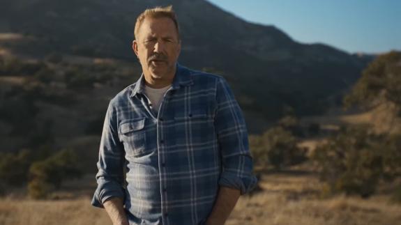 Kevin Costner stands outdoors in a sunlit, rugged landscape with rolling hills and mountains in the background, wearing a blue plaid shirt and looking toward the camera.