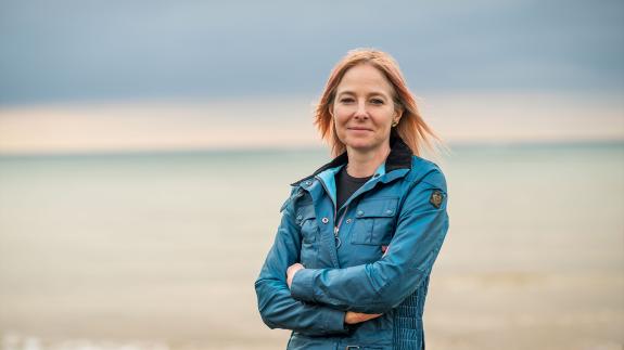 Professor Alice Roberts standing with her arms folded in front of the sea