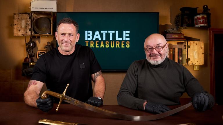 Jason Fox (L) and Bruce Crompton (R) sit in front of a television displaying the 'Battle Treasures' logo and with a large sword in front of them
