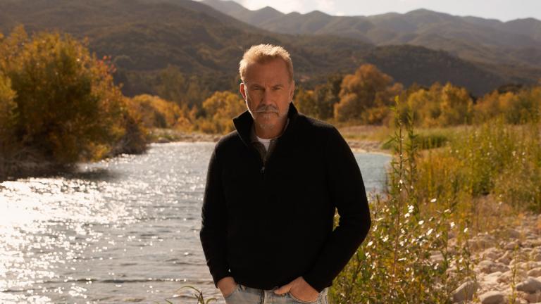 Kevin Costner stands next to a stream in the middle of rural America