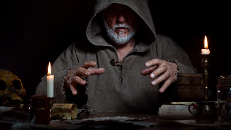 Hooded man with white beard sits at candlelit table surrounded by scrolls, bottles, and a skull