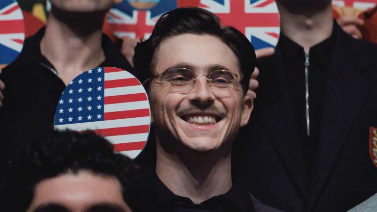 Timothée Chalamet in 'Marty Supreme' smiles while holding a ping pong paddle with the US flag on it